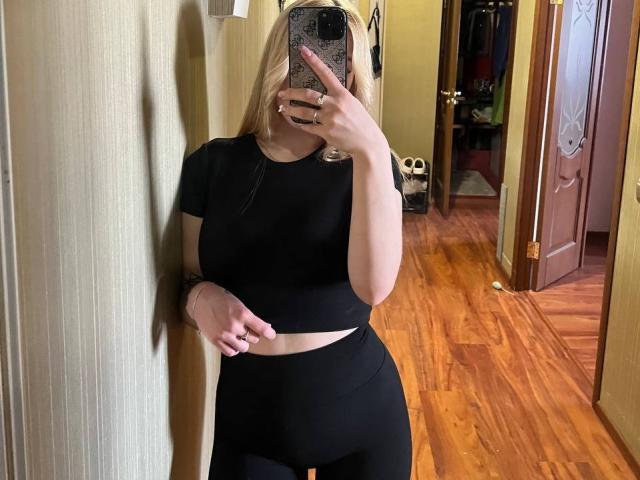 WildMaksi with blonde hair, black crop top, and black high-waist pants, holding phone in mirror selfie, showing her bare midriff and small tattoo on right wrist.