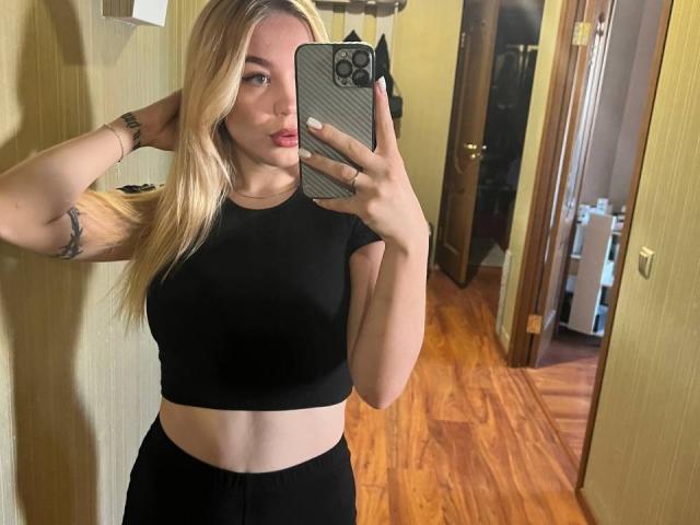 WildMaksi with blonde hair, black crop top, and black pants, taking a mirror selfie with her right hand in her hair, showing her tits and a tattoo on her left arm.