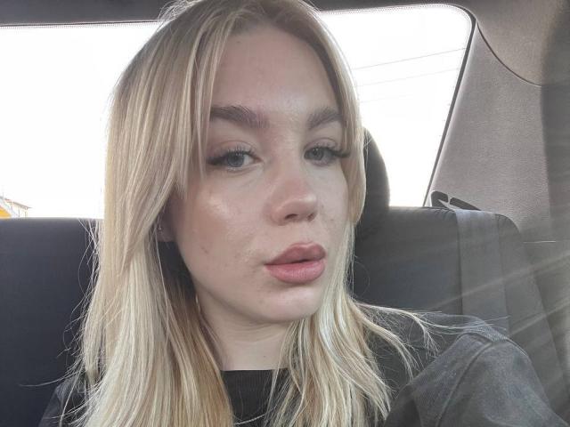 WildMaksi has blonde hair, blue eyes, and pouty pink lips. She's sitting in a car with a slightly shiny face and a black top.