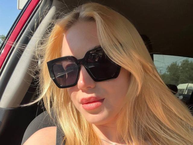 WildMaksi with blonde hair, wearing black square sunglasses, and red lipstick, sitting in a car, sunlight on her face, black tank top, slightly parted lips.