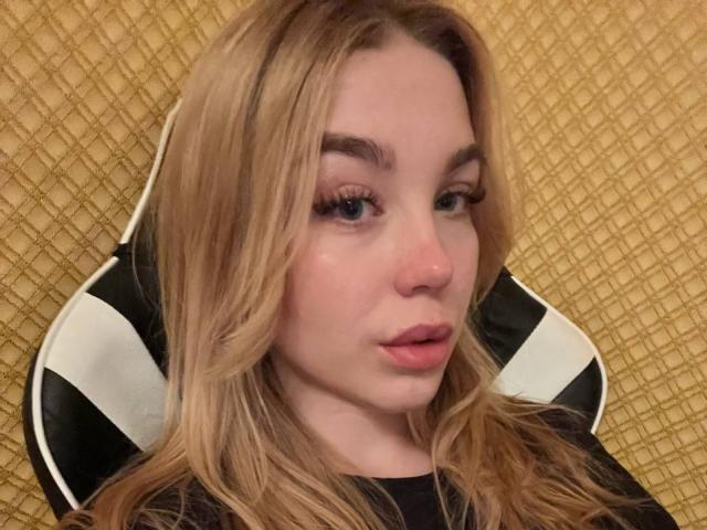 WildMaksi with blonde hair, full lips, and blue eyes, sitting in a black and white chair, wearing a black top, looking slightly to the side with a neutral expression.