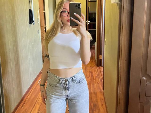 WildMaksi with blonde hair, glasses, white crop top, showing tits, and light gray jeans, taking a mirror selfie, holding an iPhone, standing in a wooden floor hallway.