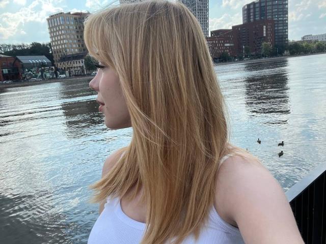 WildMaksi with blonde hair, white tank top, facing left, by a reflective waterway, showing her side profile with visible upper arms and subtle ass. Ducks swim in the background.