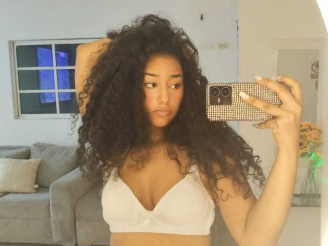 EmaWhow, avec ses cheveux noirs bouclés, portant un soutien-gorge blanc, tenant un téléphone avec une coque à carreaux, se regardant dans un miroir, montrant ses seins..