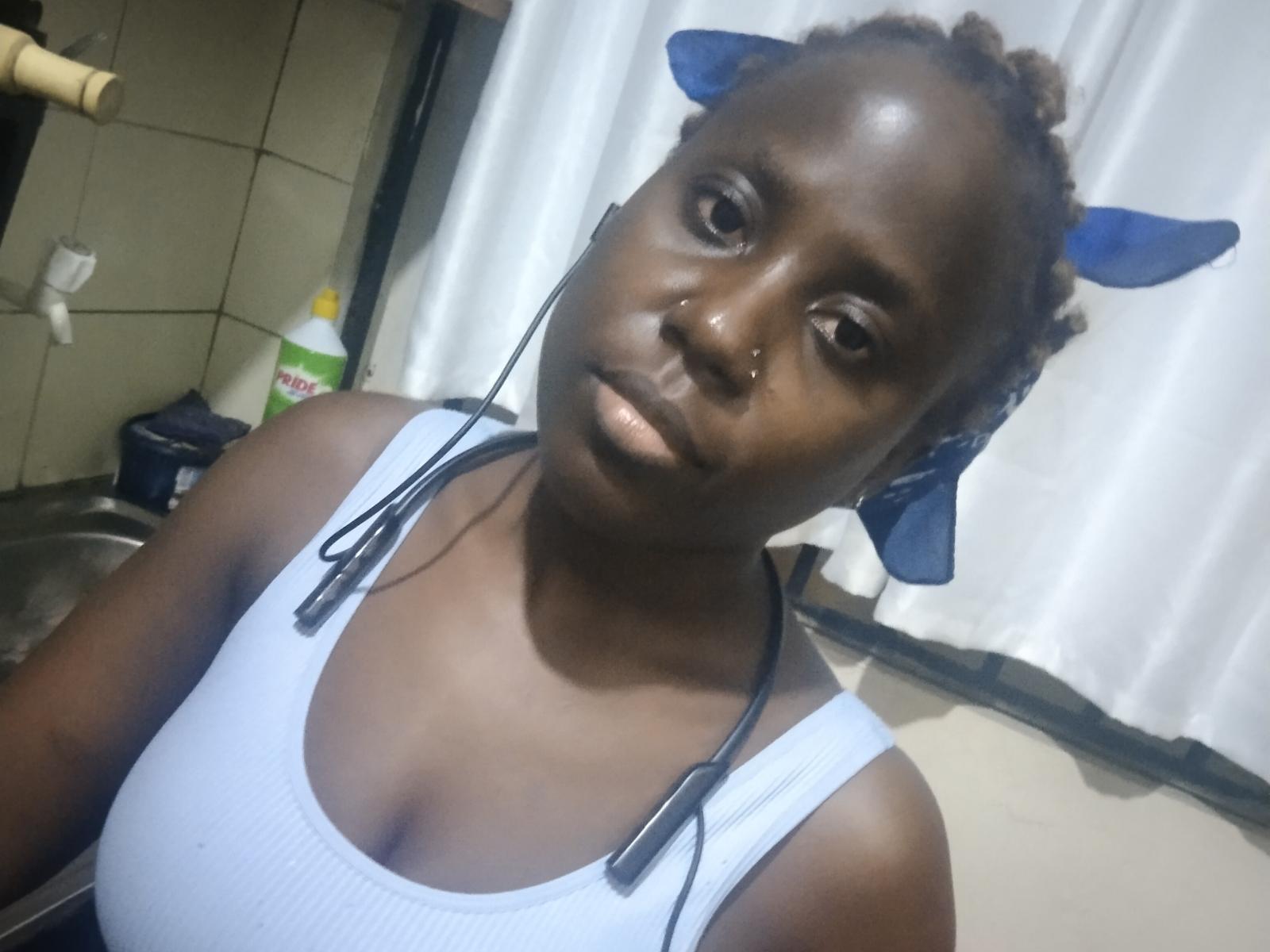 NanaBlack, a dark-skinned woman with a serious expression, wears a white ribbed tank top and has blue earbuds in. Her curly hair is tied back with blue hair ties. A small black nose piercing is visible on her right nostril. She has a stethoscope around her neck. The image is a photograph with a focus on her upper body and face.