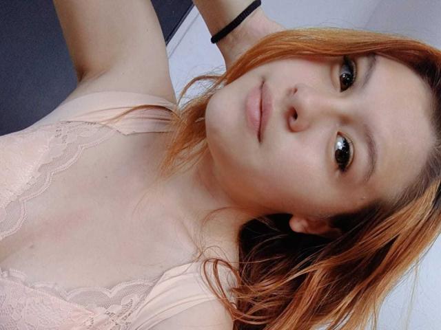 TaylorCarpert with light skin, red-orange hair, and small tits in a pink lace top, looking at the camera with a slight smile, wearing a black bracelet.