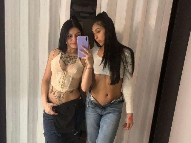 KimNKitty with black hair, tan skin, and tattoos, wearing a beige top, shows off her tits, standing next to KimNKitty with long black hair, white crop top, and blue jeans.