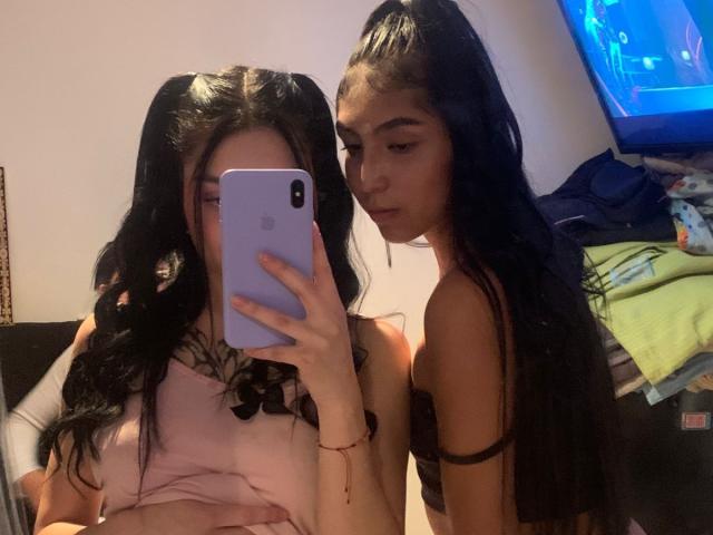 KimNKitty met zwarte vlechtjes en een roze top, die een telefoon vasthoudt, toont haar vriendin met zwarte vlechtjes en een zwarte beha, beiden kijken naar een spiegel, in een zwak verlichte kamer..