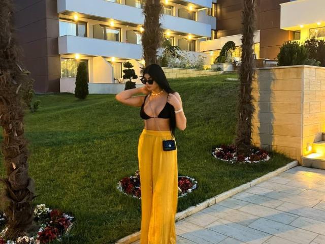 RavennaDarcy with long black hair, wearing black bra, yellow high-waisted pants, black crossbody, sunglasses, holding hair, standing in front of palm trees, lush green lawn.