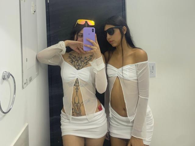 KimNKitty e un'altra donna vestite con abiti bianchi trasparenti, che mostrano tatuaggi e la parte superiore del corpo, sono in piedi in un bagno, entrambe con occhiali da sole, una delle due tiene un telefono. Seno, glutei e vulva non sono visibili..