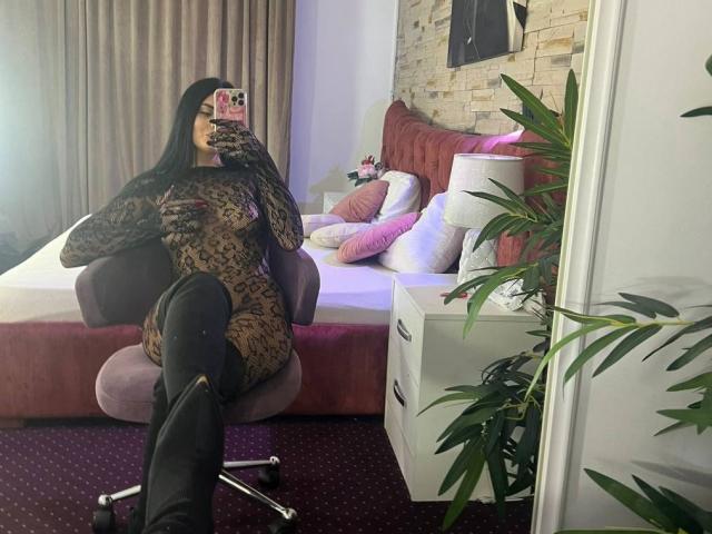 RavennaDarcy in a leopard-print bodysuit, black thigh-high boots, sitting in a pink chair, taking a mirror selfie with phone, showing her curvy ass and small tits.