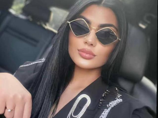 RavennaDarcy with long black hair, wearing gold hexagon sunglasses, black jacket with white patterns, and pink lipstick, looking straight at the camera.