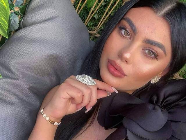 RavennaDarcy has long black hair, green eyes, and pink lips, wearing a huge diamond ring, black bow, and gold bracelet, lying on gray cushions with green leaves.