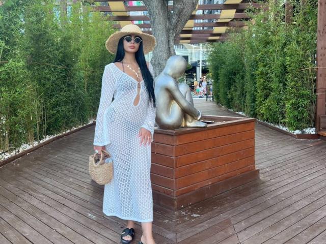 RavennaDarcy in a white, see-through dress, straw hat, and sunglasses, holding a wicker bag, standing next to a bronze statue of a sitting, faceless figure on a wooden path.