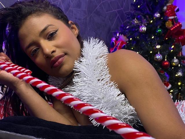 KellySmash has brown skin, dark hair, and is holding a red and white candy cane with her right hand, wearing a silver tinsel garland, and a black top, looking at camera.