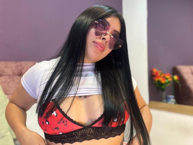 KateWelsin with long black hair, wearing pink sunglasses, a white t-shirt, and a red bra with black lace and strawberry patterns, showing her tits.