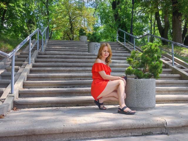 RuthPark, squatting on concrete stairs, wearing a red off-shoulder dress, black sandals, with blonde hair, showing her ass, next to a potted tree, in a sunny park.