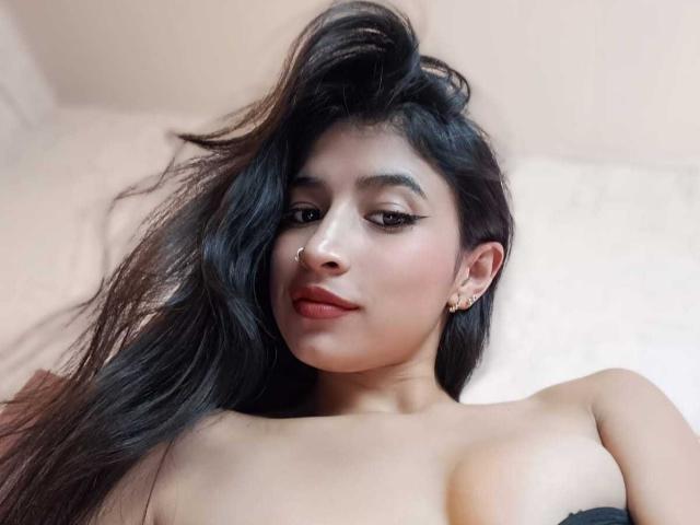 NaiaSanders with long black hair, nose ring, eyeliner, and earrings, looking down with a slight smile, showing her bare shoulders and small tits. Light skin, brown eyes.