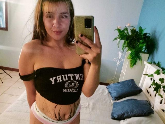 SamanthaaMiller with long brown hair, off-shoulder black top, showing her tits and a tattooed belly, wearing white panties, taking a mirror selfie with a gold phone.