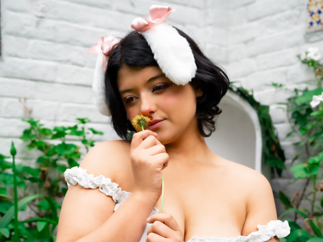 BrunnetAlba with short black hair, white bunny ears, and off-shoulder ruffle top, holding a small green plant, smiling softly, showing her tits, in a garden.