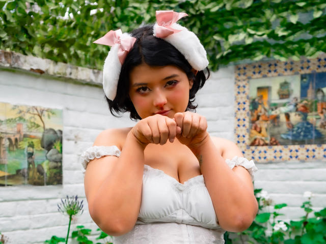 BrunnetAlba with short black hair, white off-shoulder dress, pink bow headband, hands near mouth, showing small tits, cute, green-eyed, soft smile, white brick wall in background.