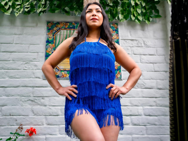 ChanelBrouw with hands on hips, wearing a blue fringed dress, showing her ass, standing against a white brick wall, long dark hair, confident expression, leafy green background.