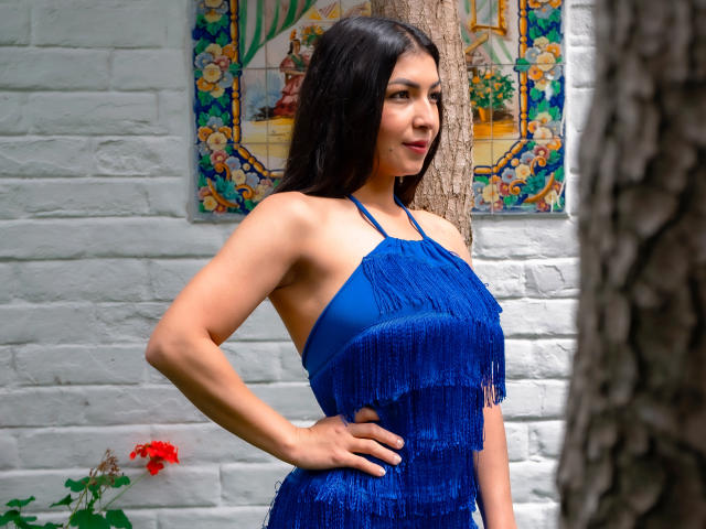 ChanelBrouw with dark hair, wearing a blue fringe dress, standing with one hand on her hip, showing her tits, against a white brick wall and tree.