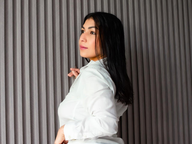 ChanelBrouw with black hair, white shirt, and pink lipstick, looking over her shoulder, showing her tits, against a ribbed gray wall.