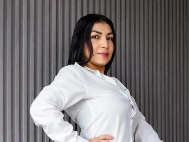 ChanelBrouw with black hair, white shirt, and pink lipstick, standing with hand on hip, showing small tits and confident smile, against ribbed gray wall.