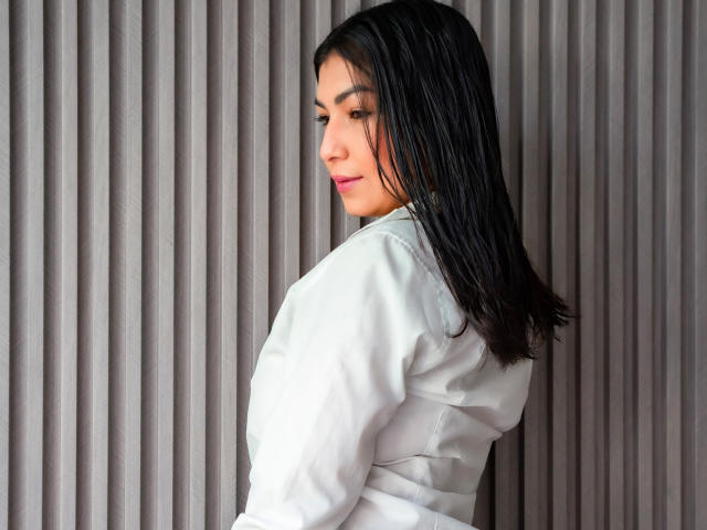 ChanelBrouw with straight black hair, wearing a white shirt, looking over her shoulder, smiling slightly, with pink lipstick, showing a small, cute butt.