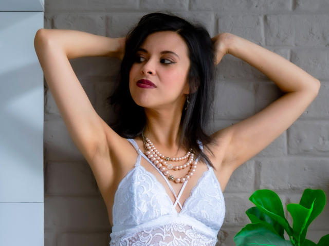 TaylaCooperk with arms raised, wearing white lace bra, pearl necklace, and red lipstick, has dark hair, fair skin, and a confident, sexy expression. Boobs visible, pussy not shown.