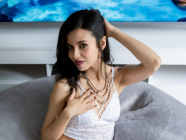 TaylaCooperk with black hair, white lace bra, pearl necklace, hand in hair, gray beanbag, blue water background, smiling, medium tits, fair skin, dark lipstick, and a sexy look.