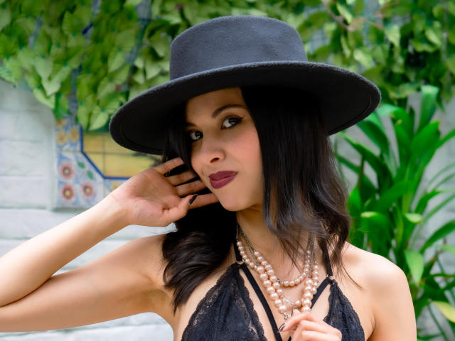 TaylaCooperk with dark hair, black hat, and pearl necklace, wearing a black lace halter, touching her cheek, looking at the camera, showing her dark red lipstick.