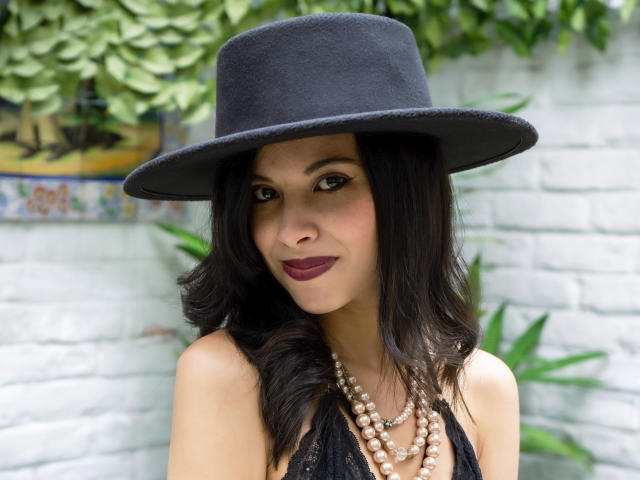 TaylaCooperk with dark wavy hair, black hat, and maroon lipstick, wearing a black lace top and pearl necklace, has a cute smile and a sexy, attractive face.