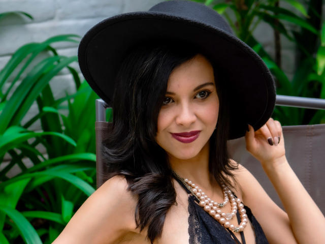 TaylaCooperk with dark wavy hair, black hat, pearl necklace, and red lipstick, holding hat, showing cleavage in black lace top, smiling confidently.
