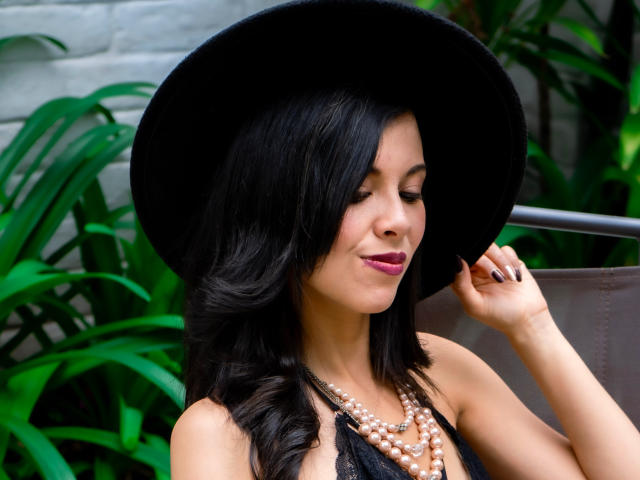 TaylaCooperk with black hair, wearing a big black hat, pearl necklace, and pink lipstick, smiling, holding her hat, showing her boobs, and looking down.