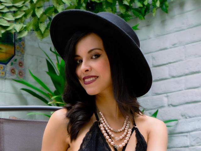 TaylaCooperk with dark wavy hair, black hat, black lace halter, pearl necklace, and purple lipstick, smiling at camera, showing off her tits. Green plant in background.