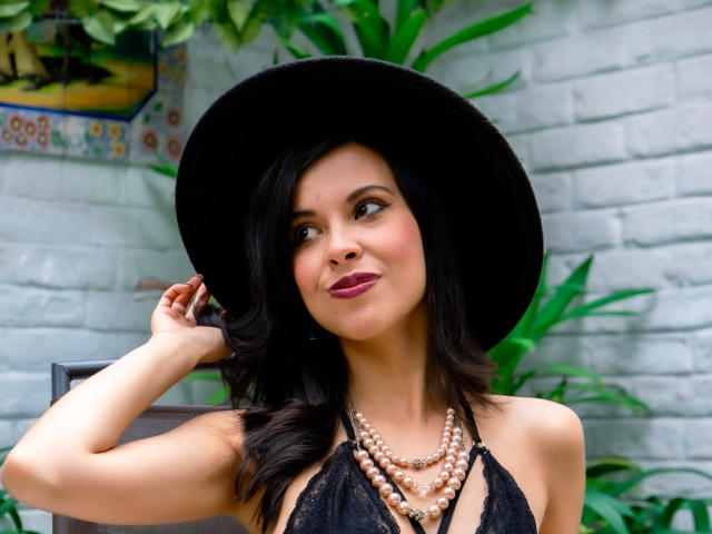 TaylaCooperk with black wavy hair, wearing a black hat, black lace halter top, and pearl necklace, looking up with a slight smile, showing her boobs.