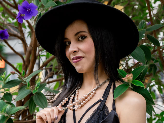 TaylaCooperk has black hair, wearing a black hat, pearl necklace, and black lace top, with purple flowers in the background. She's looking at the camera with a slight smile.