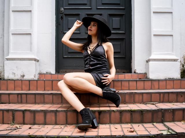 TaylaCooperk sits on brick steps, legs crossed, wearing a black hat, shiny black dress, and black boots, hand touching hat, showing off her ass and smooth legs.