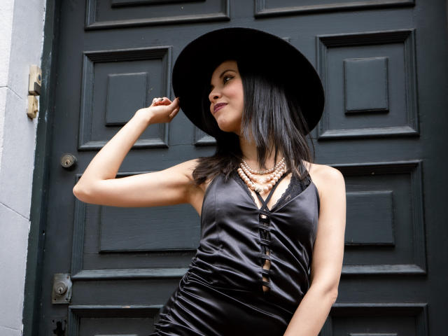 TaylaCooperk with black hat, shiny black dress, and beads, showing off her tits and ass, standing in front of a dark door, hand on hat, looking to the side.
