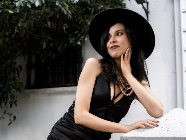 TaylaCooperk with black dress, big tits, black hat, purple lipstick, and necklace, leaning on white wall, hand on cheek, looking up, dark hair, green leaves in background.