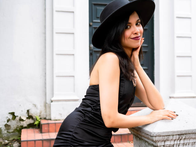 TaylaCooperk with black hat, black dress, and bare shoulders, leaning on a stone ledge, showing off her ass, smiling, with dark nails and a subtle makeup look.