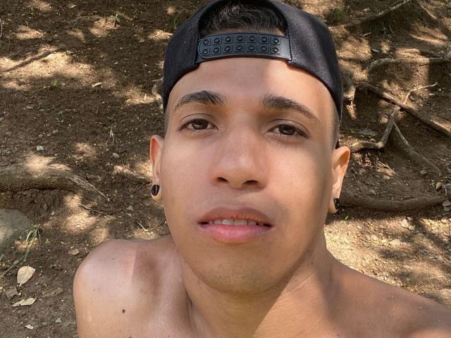 Yoximanuelprime with a shirtless face, dark eyebrows, black backward cap, ear plugs, slightly open mouth, and a tan, dirt-covered background. His brown eyes and short dark hair are visible.