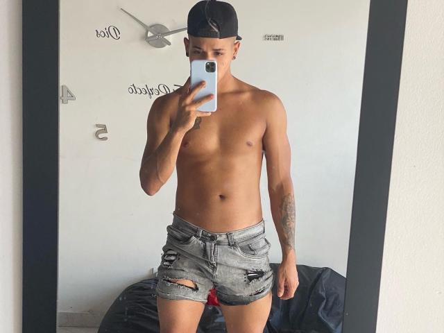 Yoximanuelprime, ohne Hemd, mit schlanker, trainierter Figur, trägt zerrissene Jeansshorts, eine schwarze Kappe und hält ein Telefon. Sein Spiegelbild zeigt ein Tattoo auf seinem rechten Arm..