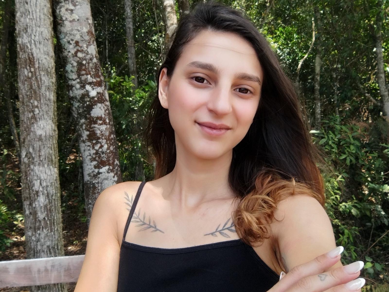 DiabaSelvagem, a cute woman with brown hair, small tits, black tattoo on chest, smiling, wearing black top, holding arm, forest background, natural light, white nails, slight tan.
