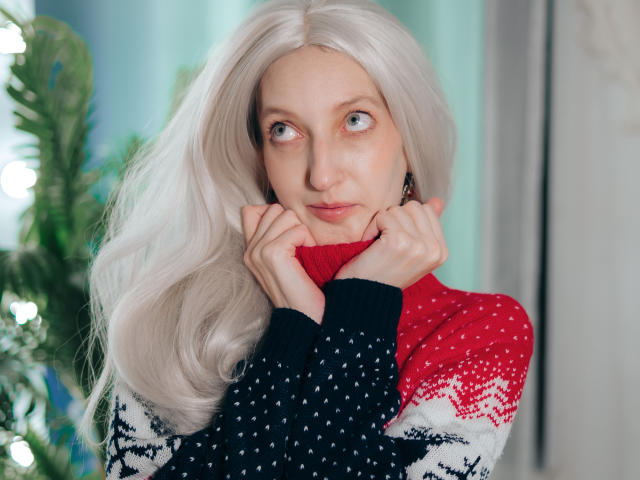 ViktoriaClark with long white hair, wearing a red and black patterned sweater, holding her chin with her hands, looking up with a cute, slightly curious expression.