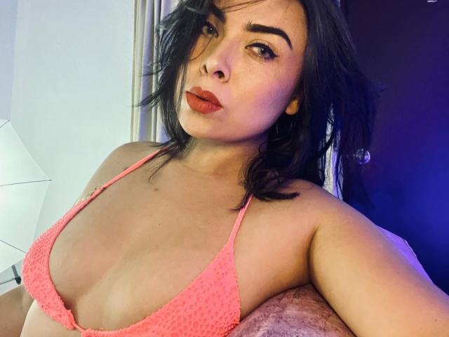 CataGuerra with dark hair, orange lipstick, and pink, textured bikini top, showing small tits and a pouty face, looking into the camera. Her eyebrows are thick and she's sexy.