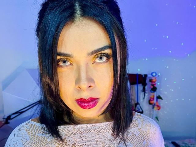 CataGuerra has dark hair, red lipstick, and is wearing a white lace top. Her eyes are wide and she's staring directly at the camera with a slight pout.