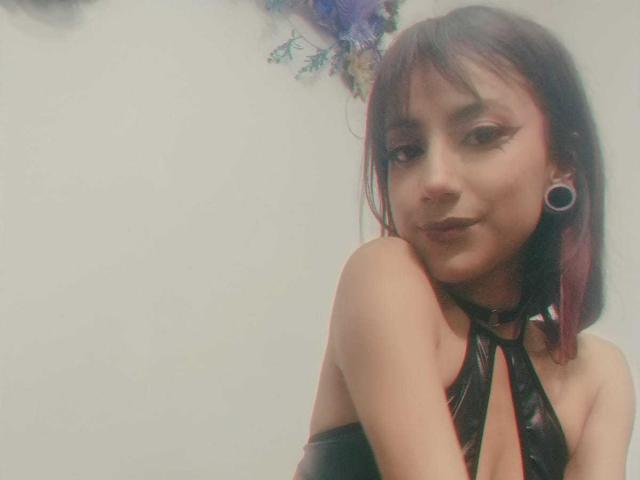 KarlaMiilan, avec ses cheveux roux foncés, son eye-liner noir et son anneau de nez, porte un haut en cuir noir à encolure dégagée, laissant entrevoir ses seins, et sourit à l'appareil photo. Elle a des décorations florales violettes..